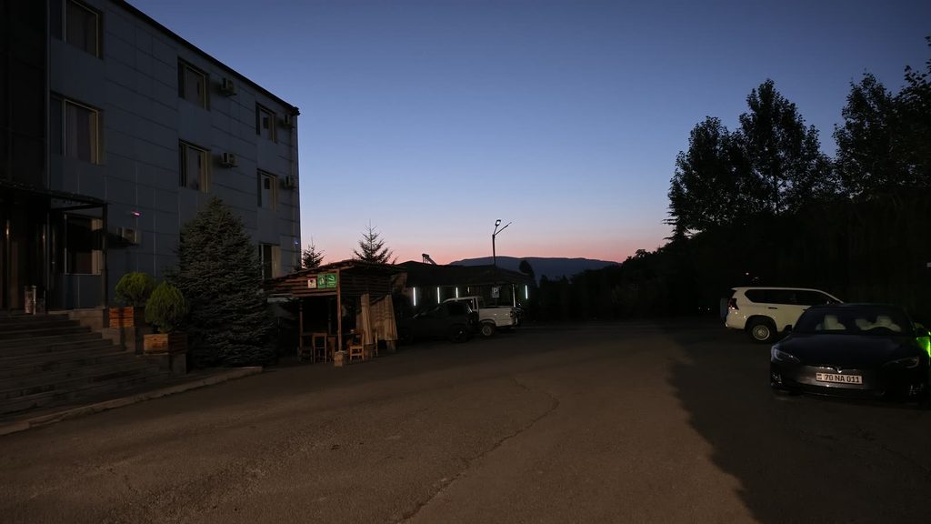 A dark four-storey hotel building on the left and parked cars on the right, under a deep blue sky with a band of orange and pink light along the mountains on the horizon.