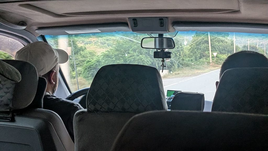 The inside of a minibus seen from the back seat: the driver in a beige cap, the headrests of the seats in front, and through the windscreen a road between green hills.