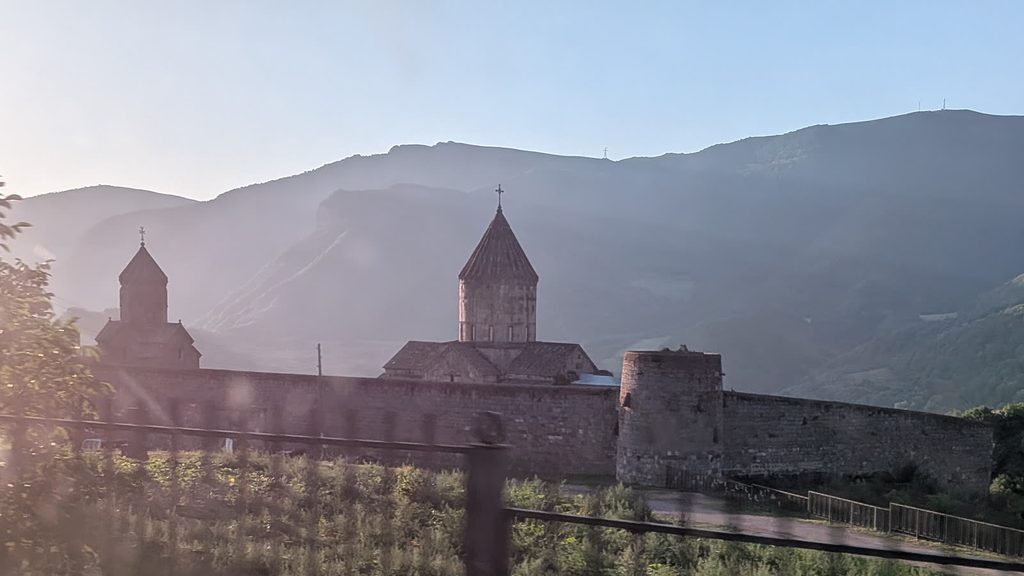 Tatev monastery in the low morning sun: a church with a conical dome and a smaller belfry behind a long stone wall with a round tower, below hazy mountain ridges, seen through a car window.