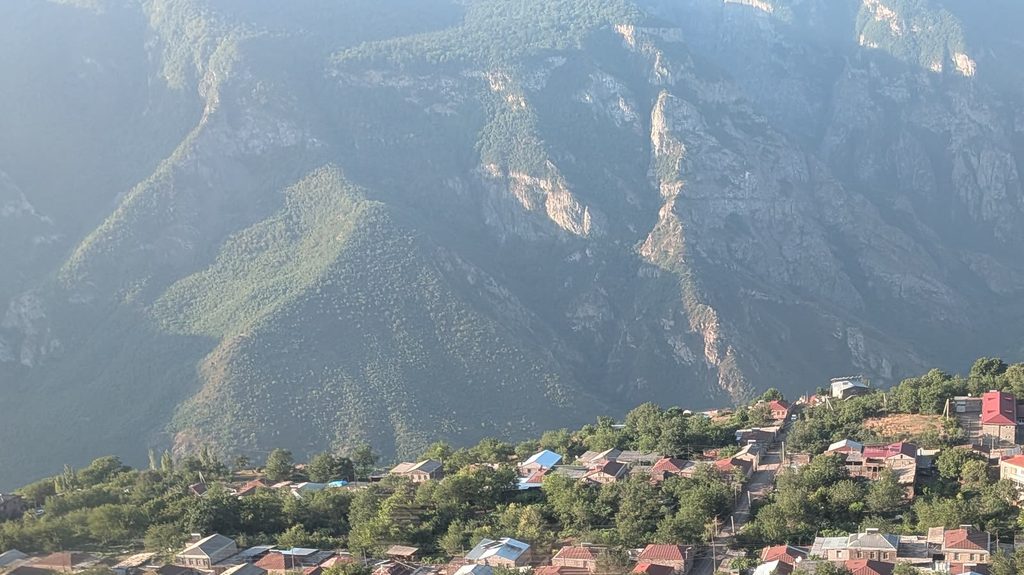 Red and blue roofs of a village among trees on a shelf of land, with the steep, wooded cliffs of a gorge rising behind it.