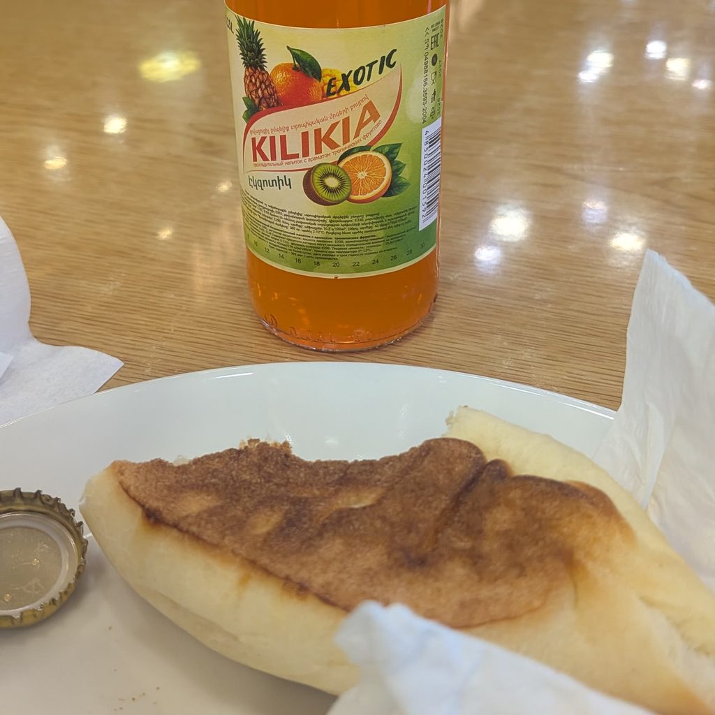 A bottle of orange Kilikia Exotic soda on a table, with a pastry on a white plate and the bottle cap beside it.