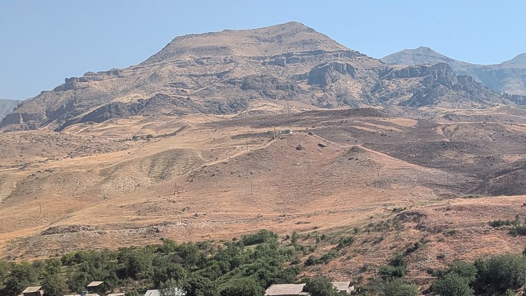 Dry golden hills and a bare, rocky mountain under a pale blue sky, above a strip of green trees and village roofs.