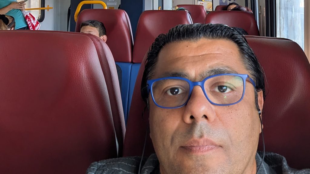 Garo Kotchounian in blue glasses taking a selfie in a train carriage with dark red seats, with other passengers sitting behind him.