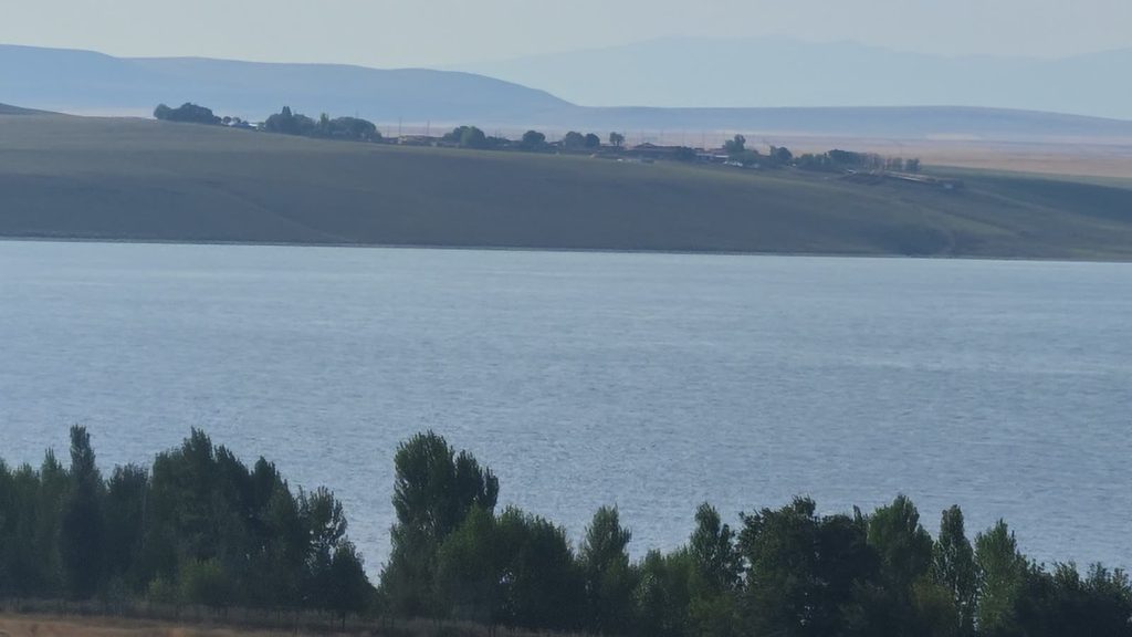 A village among trees on a green slope across a wide stretch of water, seen over the tops of poplar trees.