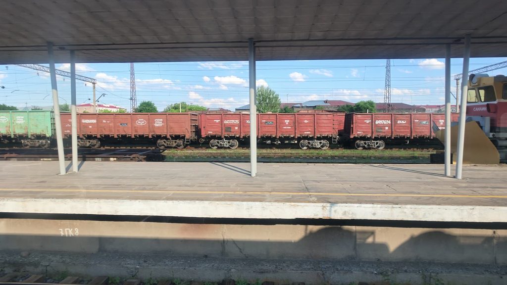 Gyumri station seen from the train: a long platform under a canopy, with a row of red freight wagons and a locomotive on the tracks beyond.