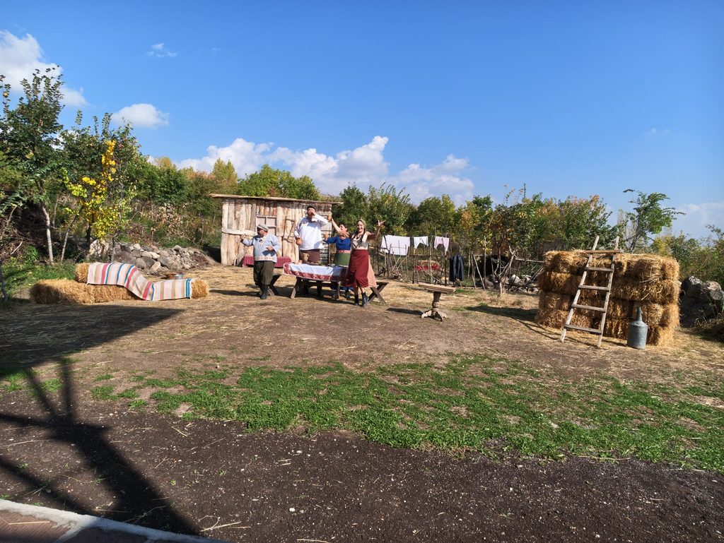 A wide view of an open-air theatre set across a grassy yard: a log shed, a striped rug over hay bales, a long table with actors around it and a haystack with a ladder, under a blue sky with white clouds.