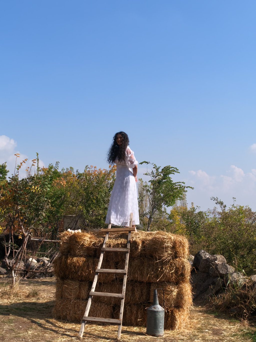 A young woman with long dark curly hair, in a white dress, stands on top of a stack of hay bales, with a wooden ladder and a metal can at its foot.