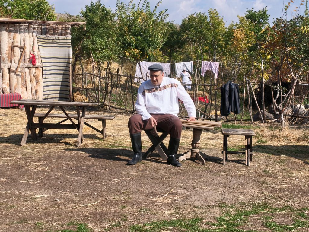 A man in a flat cap and a white shirt with an embroidered band sits on a folding stool beside a small table, with a fence of branches and a line of washing behind him.