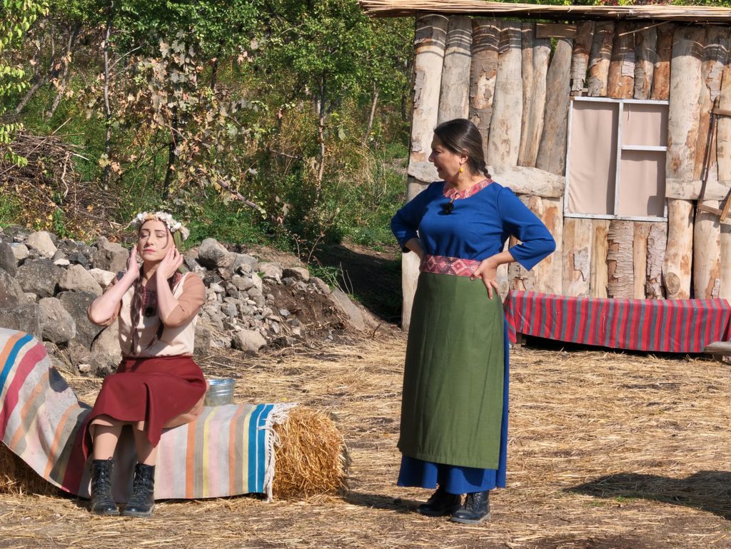 Two women in village costume: one in a red skirt and a flower wreath sits on a striped rug over hay bales with her hands to her face, while the other, in a blue blouse and a long green apron, stands with her hands on her hips in front of a log shed.