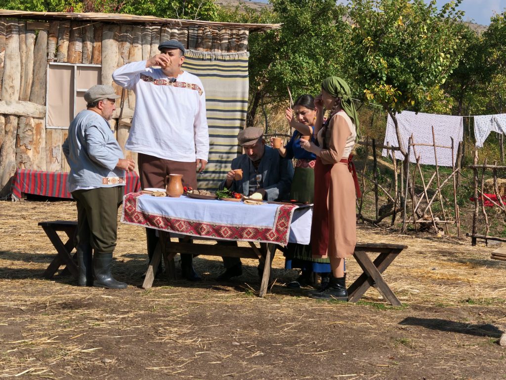 Villagers in caps and embroidered shirts stand and sit around a table laid with an embroidered cloth and a jug, in front of a log shed and a line of washing; a man in a white shirt drinks from a cup.