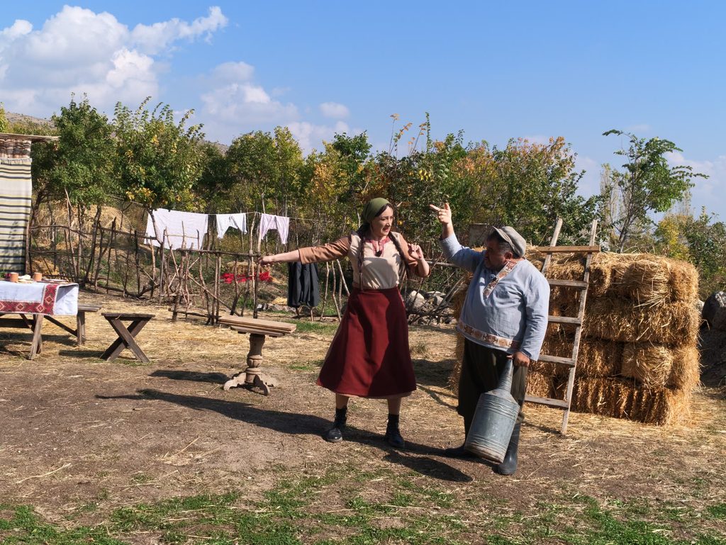 A woman in a red skirt and a headscarf spreads her arms beside a man in a cap who holds a metal can, in front of a haystack and a line of washing.