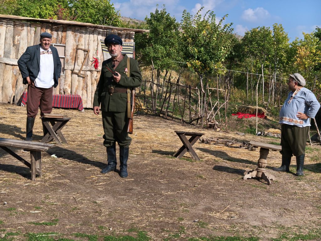 An actor in a dark green military uniform and a dark peaked cap stands in the yard between a man in a dark jacket and a man in a pale blue tunic, in front of the log shed.