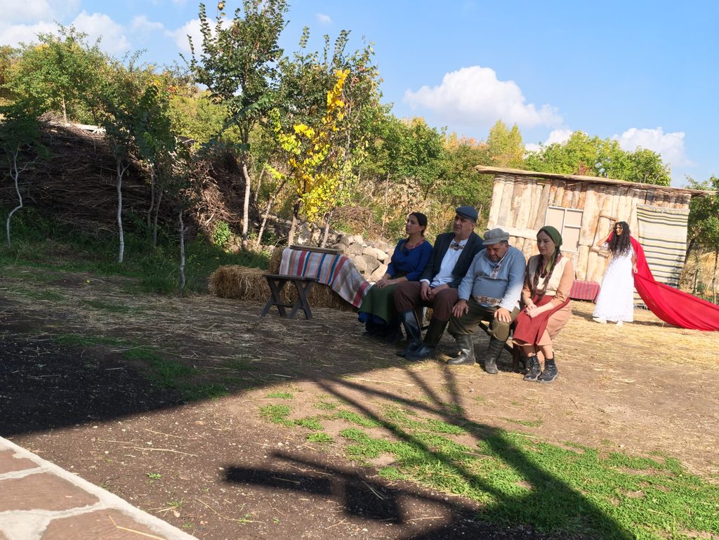 Several actors sit on a long bench at the edge of the yard while the bride, in white, stands a little apart with a long red cloth trailing on the ground.