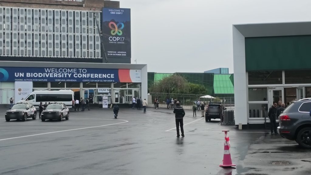 The entrance of the Meridian Expo in Yerevan on a grey, wet day, under a long blue banner reading Welcome to UN Biodiversity Conference and a tall COP17 banner, with two police cars and a white minibus in the forecourt.