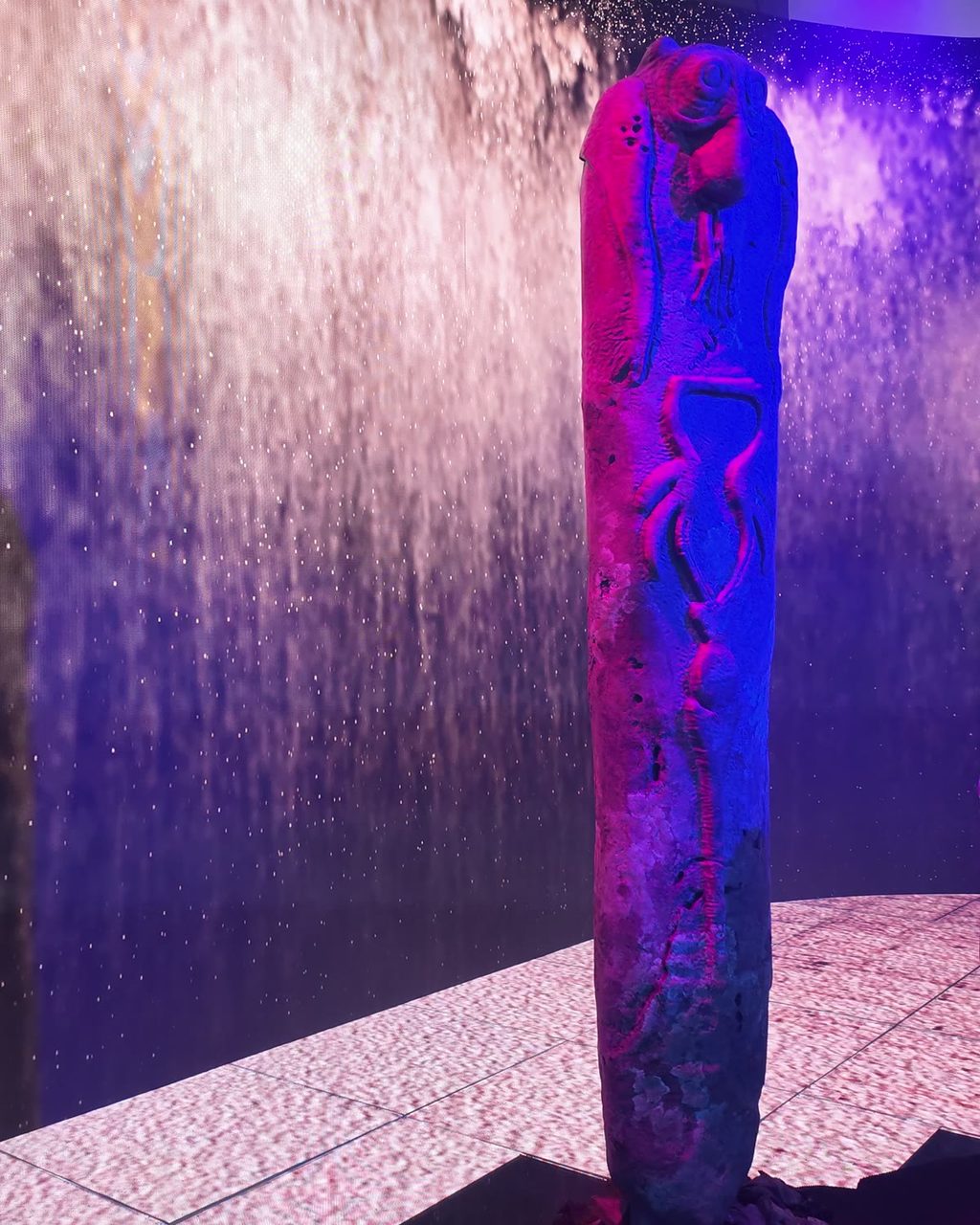 A tall carved stone standing upright in purple and blue light, in front of a waterfall projected on the wall, on a floor of glowing pale square panels.