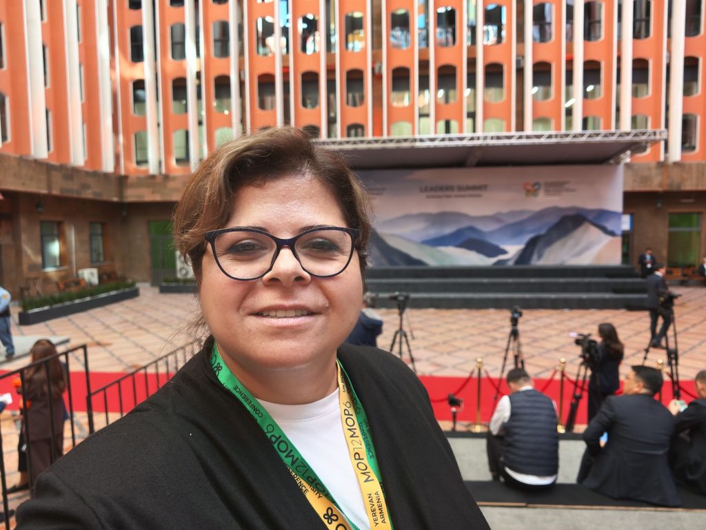 Araz, in glasses and a black cardigan with two COP17 lanyards, one green and one yellow, smiling at the camera in a paved courtyard, with a stage under a painted mountain backdrop, television cameras on tripods and an orange building behind her.