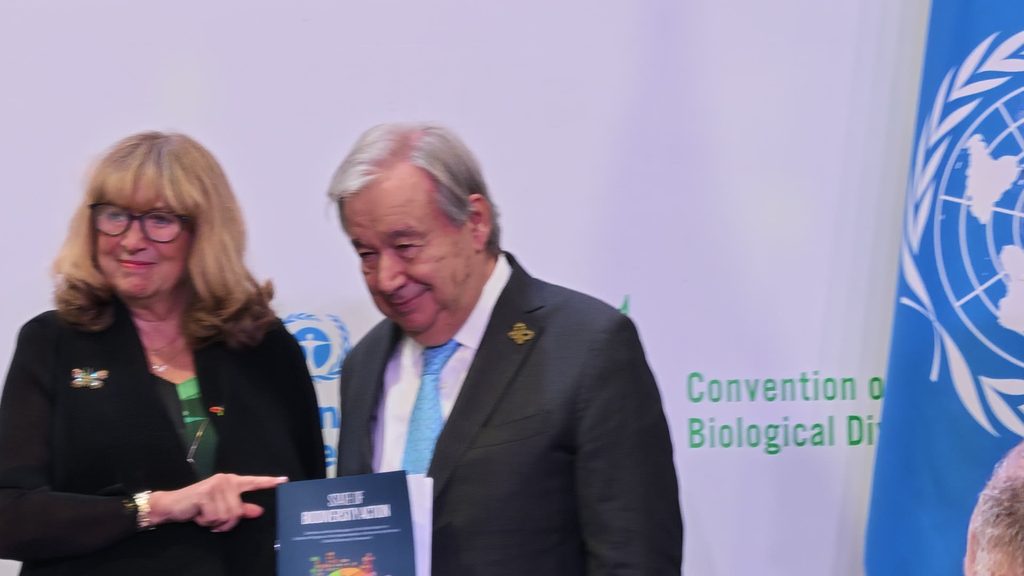 António Guterres holding a dark blue copy of the report as a woman in glasses points to it, in front of a white backdrop with the green words of the Convention on Biological Diversity, partly hidden by a blue UN flag.