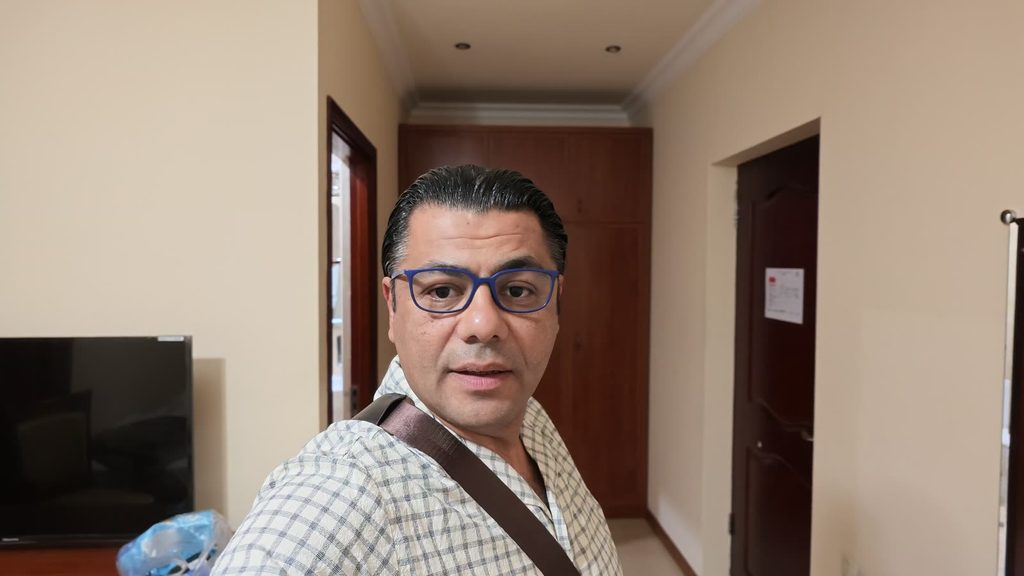 Garo Kotchounian in blue glasses and a checked shirt, a leather strap across his chest, in the cream-walled entrance of a hotel room, with a television on the left and a wooden wardrobe behind him.