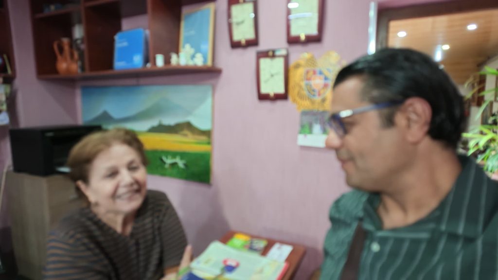 A smiling older woman at a desk talks with a man in glasses, beneath a painting of Mount Ararat and the Armenian coat of arms.