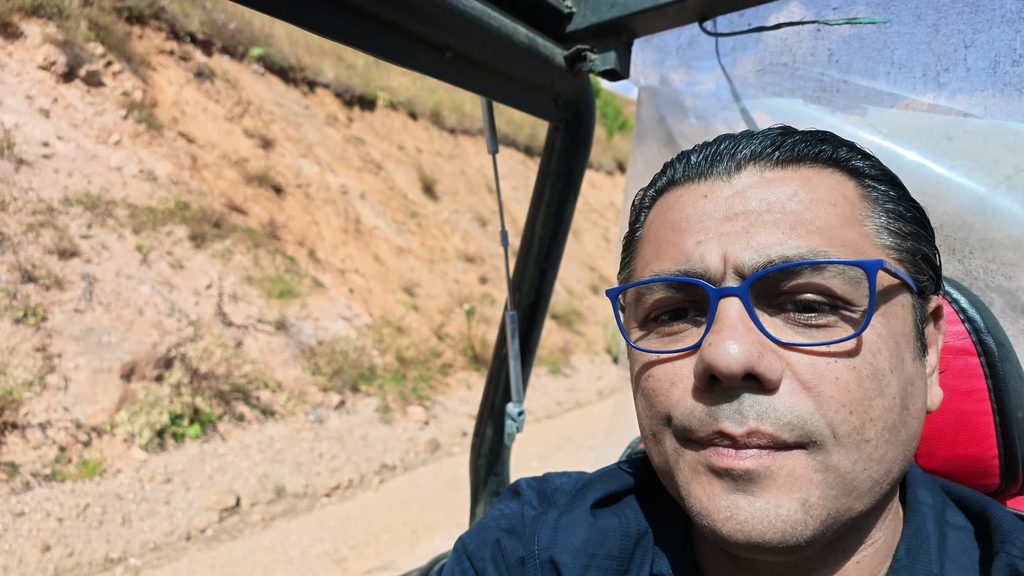 Garo Kotchounian in blue glasses, sitting in the open back of a four-by-four on a dusty mountain track, with a red seat, a black roll bar and a clear plastic screen behind him.