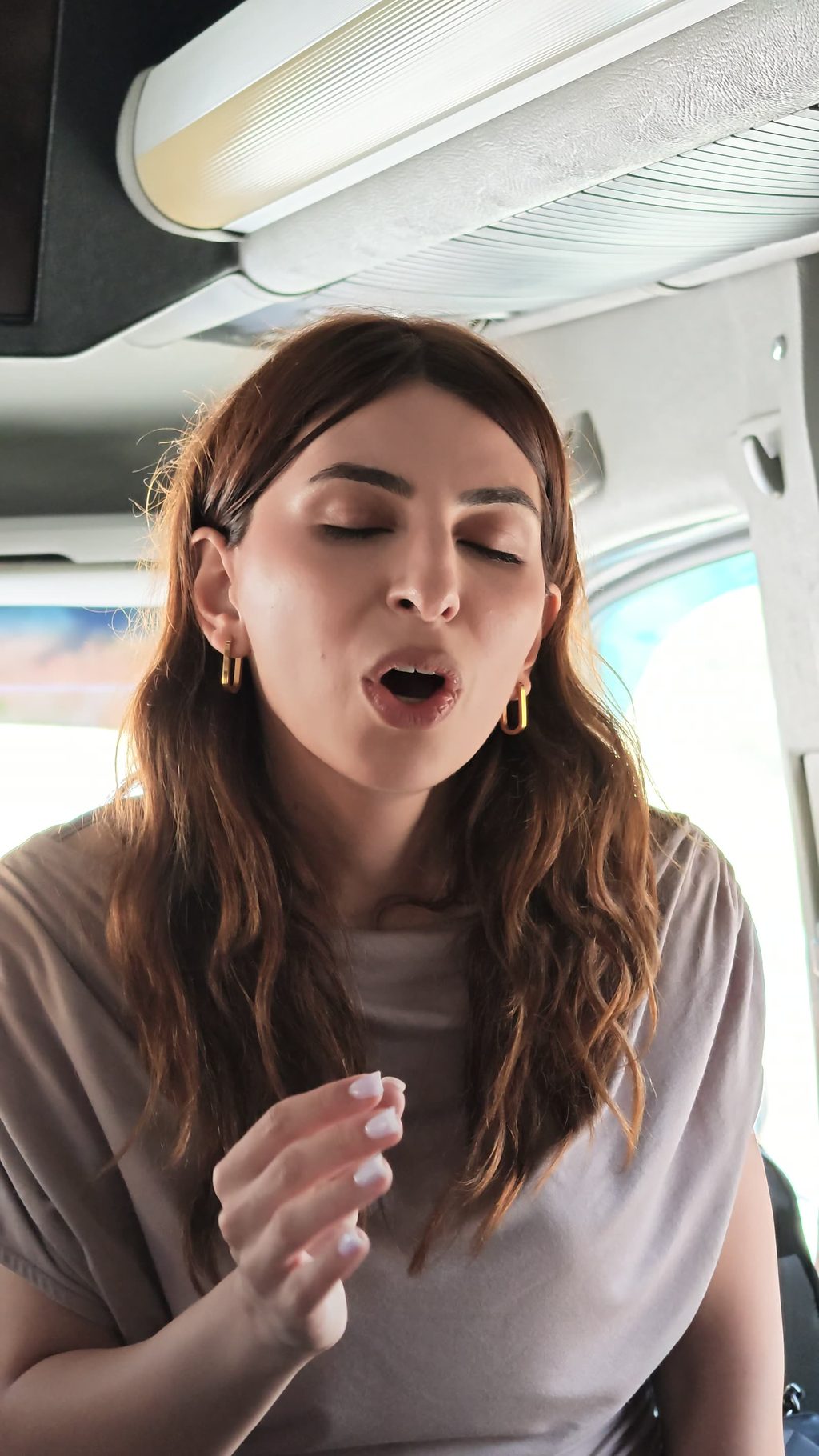 Karina singing in the aisle of the bus, one hand on a seat-back, eyes closed.