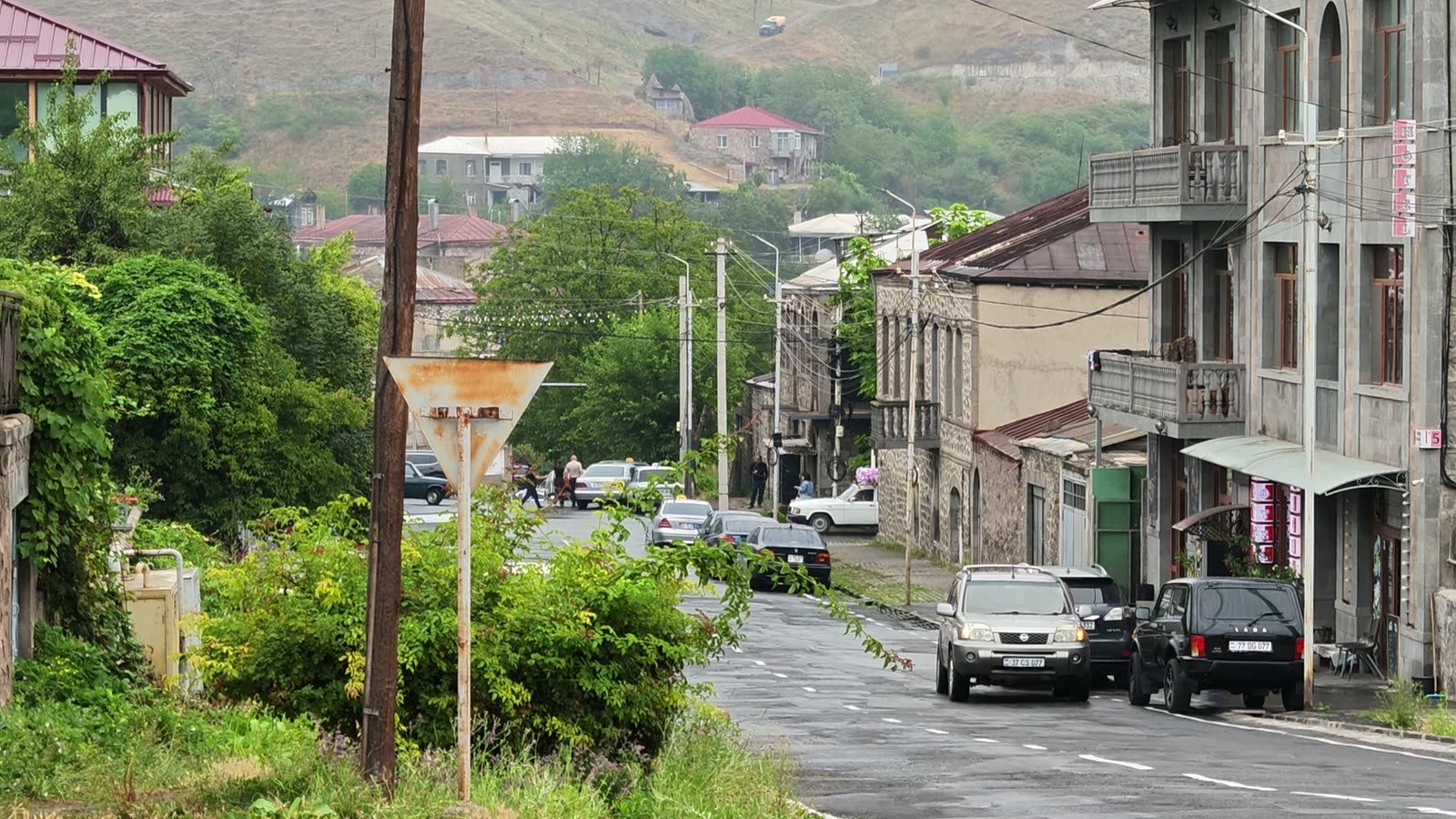 A wet street in Goris: stone houses with balconies, parked cars, and the green hillside behind.