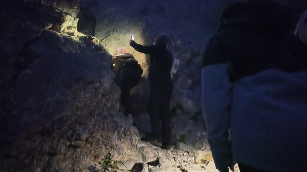 Climbers in dark clothes scrambling up a rock face at night, one of them holding up a phone whose torch lights the rock, with a figure in a pale blue fleece in the foreground.