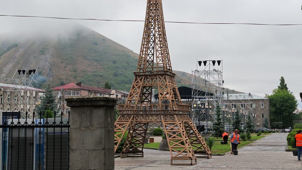 A tall lattice replica of the Eiffel Tower stands in a paved town park, with stage scaffolding and a mist-covered mountain behind it.