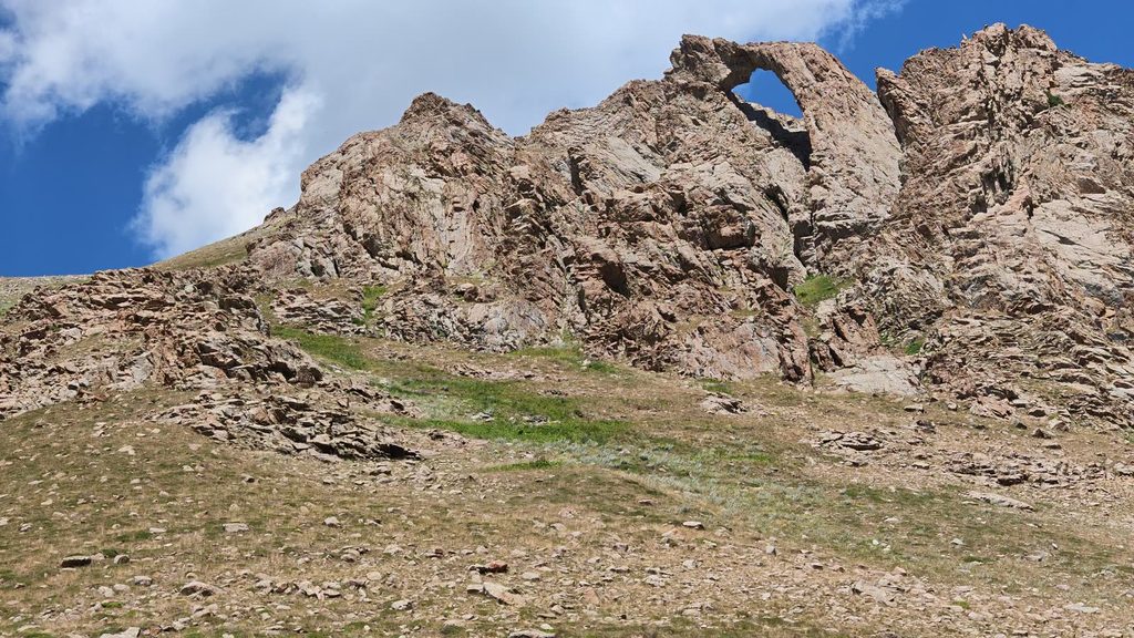 A ridge of rough brown rock with a large hole right through it, under a blue sky with white cloud, above a stony, grassy slope.