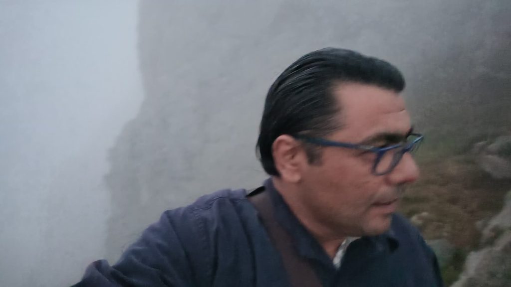 Garo in glasses and a dark jacket on the summit ridge, with grey cliffs disappearing into thick fog behind him.
