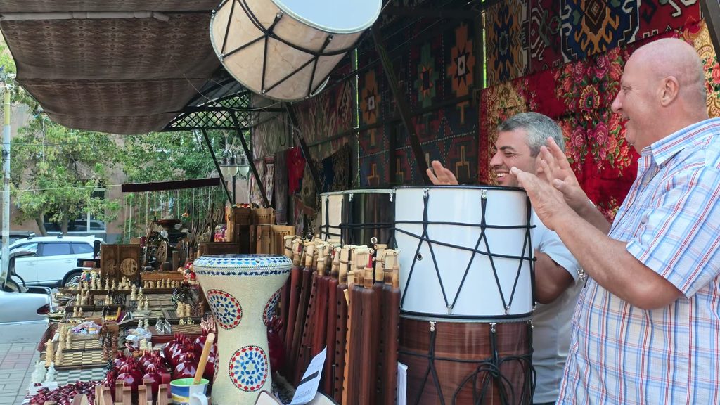 A man with a dhol drum at a market stall, mid-beat.