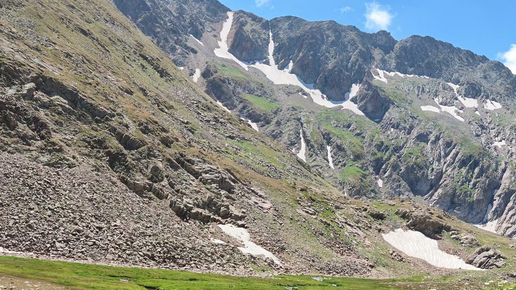 A steep grey mountainside streaked with snow in its gullies, above a scree slope and a strip of green meadow.