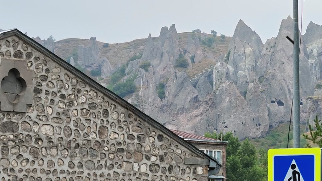 Tall grey rock spires with cave openings rise behind the fieldstone gable of a building in Goris.
