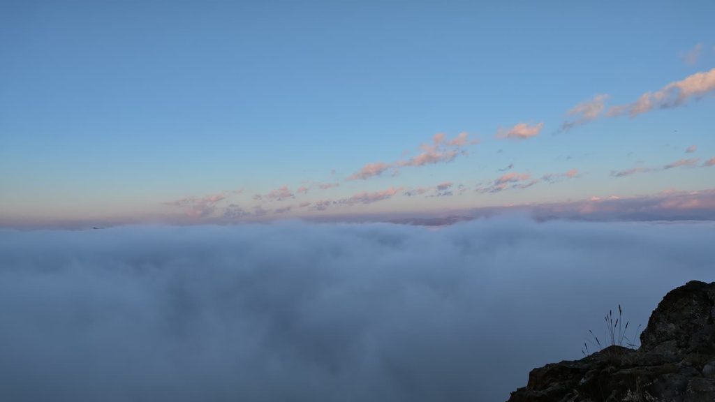 A flat sea of cloud stretching to the horizon at dawn under a blue sky with pink-lit streaks of high cloud, a dark rock in the lower right corner.