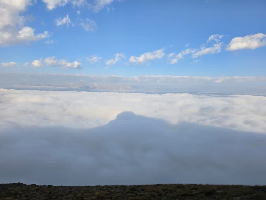 The shadow of the mountain cast as a dark triangle on a flat layer of white cloud, with a faint halo of light at its peak, under a blue sky with small clouds.