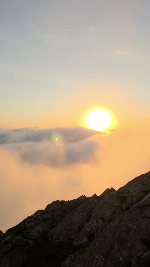 The sun rising orange above a bank of cloud, seen past a dark rocky slope.