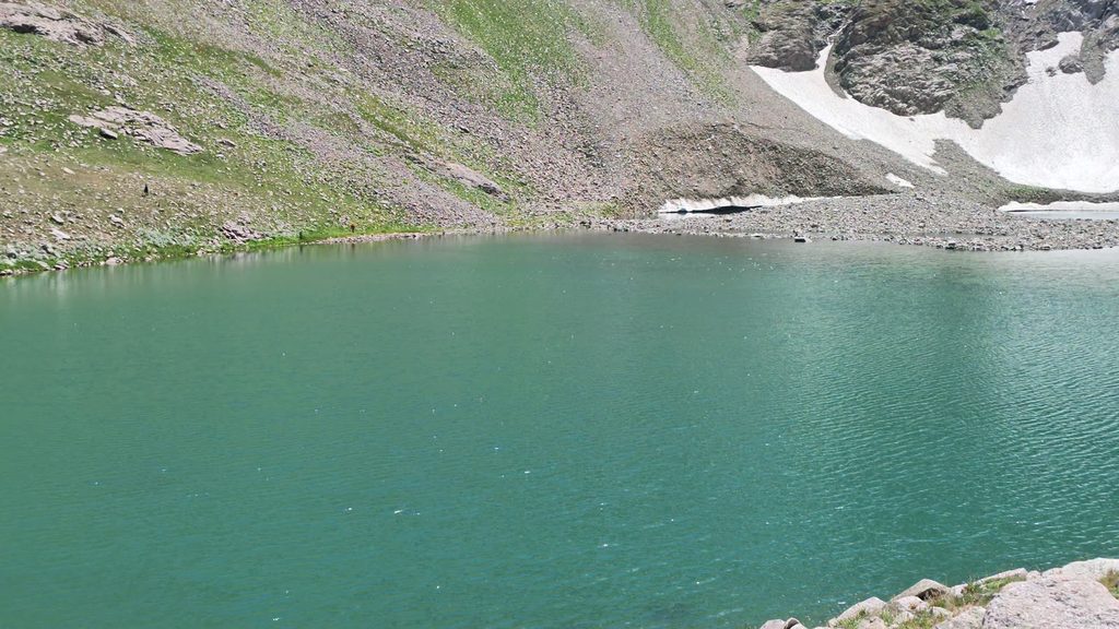 A turquoise mountain lake below a scree slope, with patches of snow along its far shore.