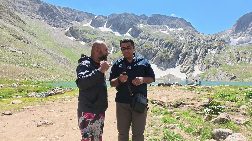 Harut, bald and bearded, in a dark jacket and patterned shorts, beside Garo, who looks at his phone, on the shore above the lake with snowy peaks behind them.