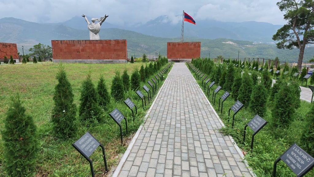 An avenue of young trees running down the hill, a plaque set in the grass at each one.