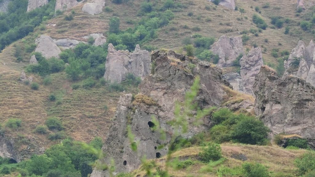 Weathered grey rock pinnacles on a grassy slope, one pierced by small dark cave dwellings.