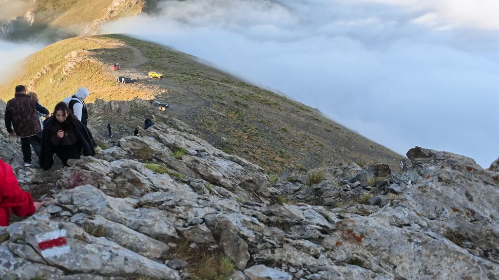 Climbers descending a rocky ridge in the morning sun, with a sea of cloud beyond and cars as small dots on the grassy plateau below.