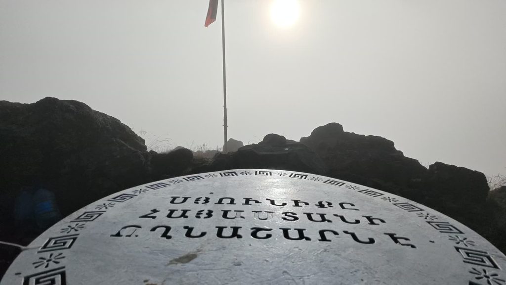 A round white plaque with a Greek-key border and the Armenian words ՍՅՈՒՆԻՔԸ ՀԱՅԱՍՏԱՆԻ ՈՂՆԱՇԱՐՆ Է in black capitals, below a flagpole and a hazy sun.