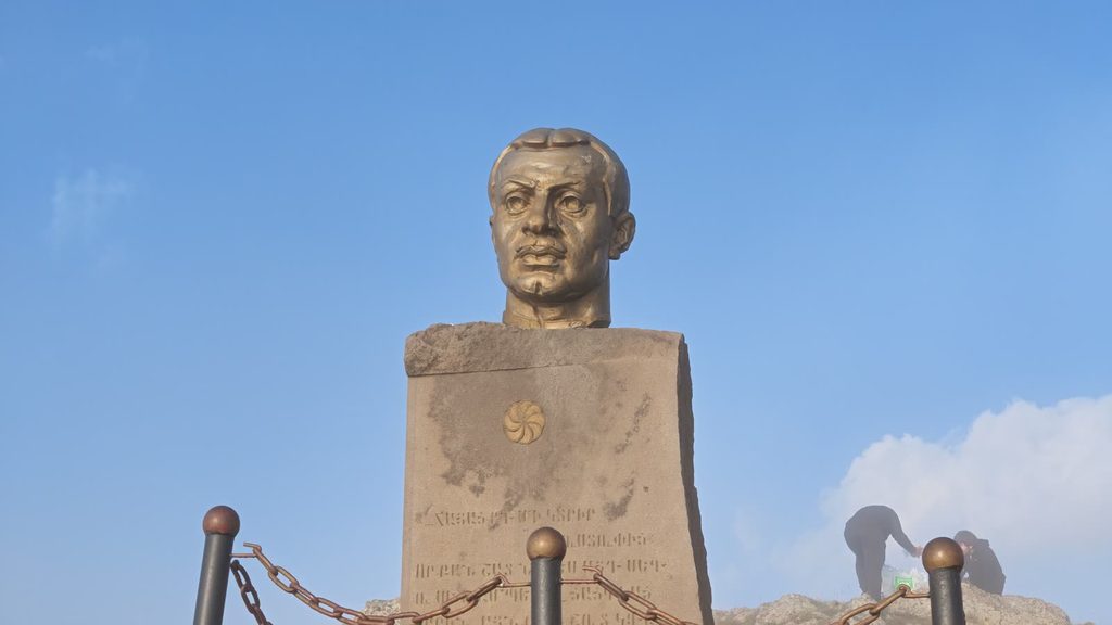 A bronze bust of Garegin Nzhdeh on a tall stone pedestal carved with a quotation and a gilt eternity sign, inside a low chain fence, against a blue sky.