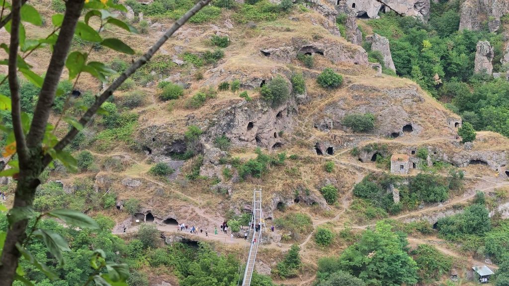 A rocky hillside riddled with carved cave dwellings and footpaths, with a small stone hut and the white tower of a suspension bridge where a line of visitors is walking.