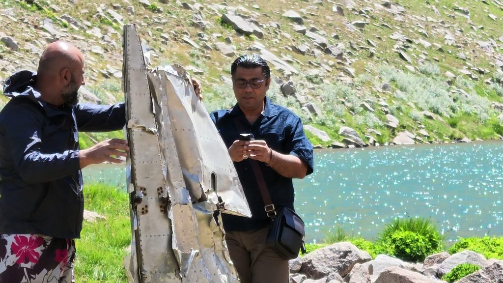 Harut holds up a long, battered sheet of riveted grey metal beside the lake while Garo looks at his phone, with the water glittering behind them.