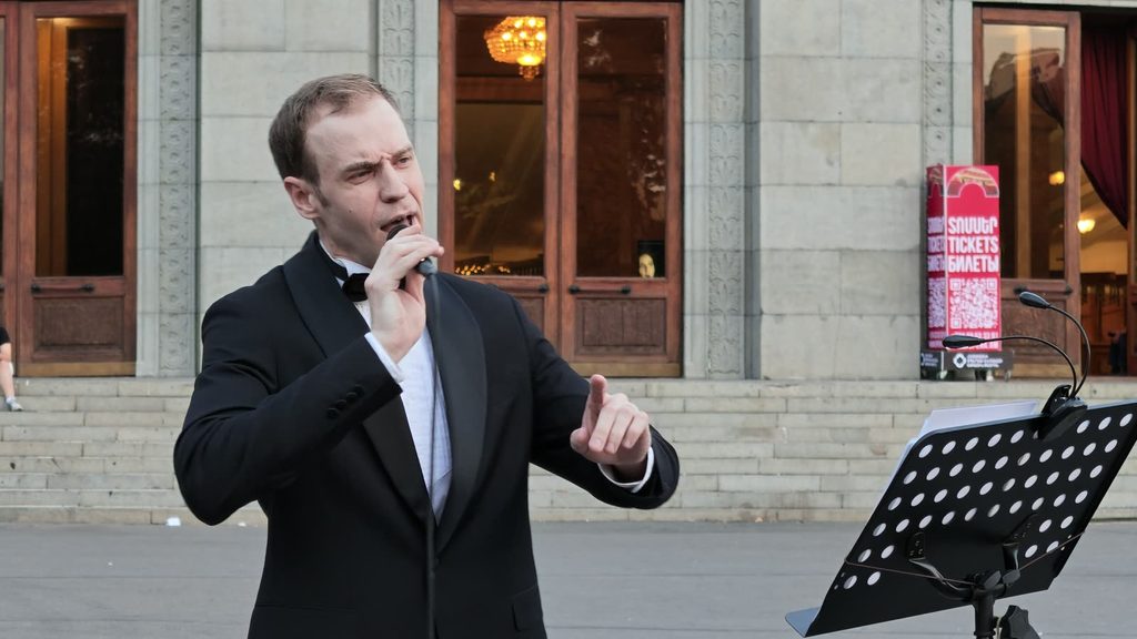 A man in full black tie singing beside a music stand in front of the Opera House.