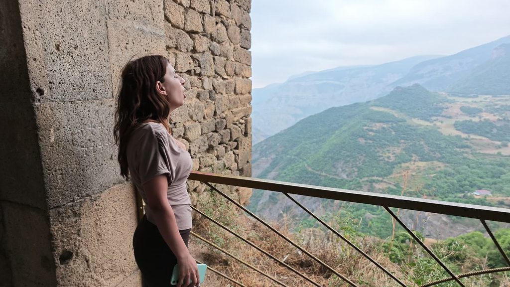 Karina singing on a stone balcony at the edge of Tatev monastery, the gorge behind her, people filming on their phones.