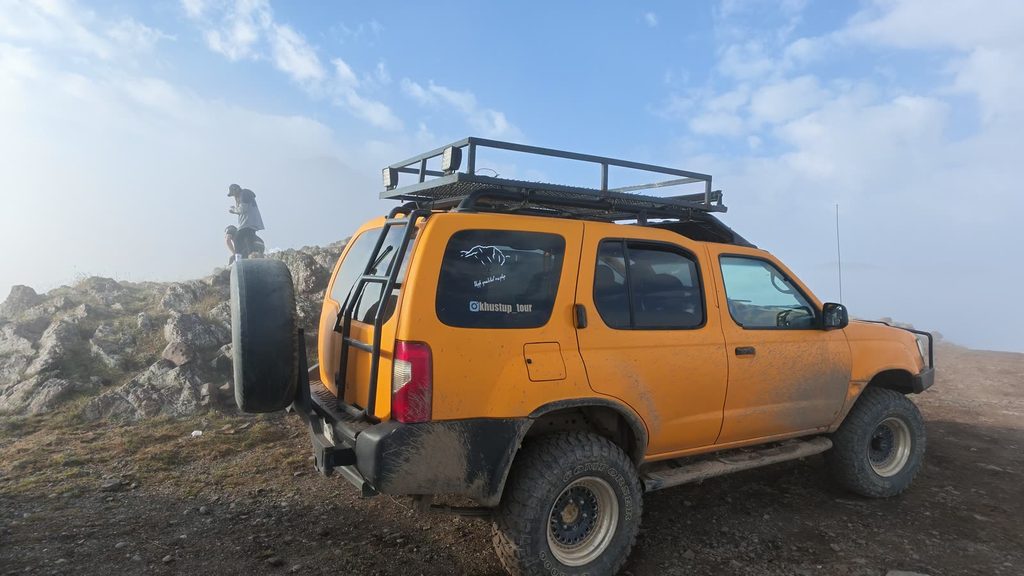 A mud-splashed yellow four-by-four with a roof rack parked on a rocky plateau, with mist and blue sky behind it.