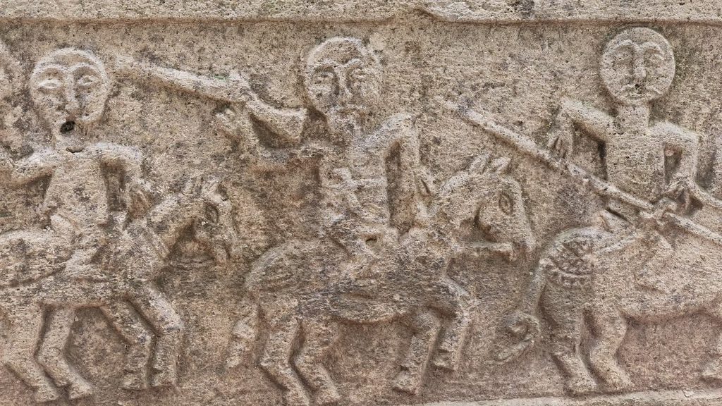 A weathered stone relief of three horsemen with round, moustached faces riding in a row, carved along the side of a tombstone.