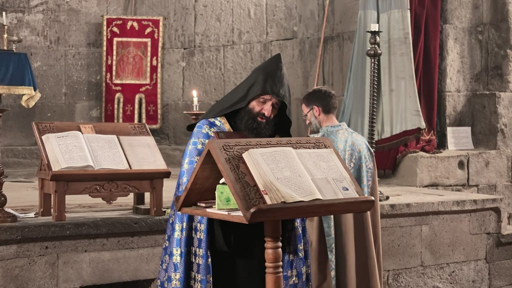 An evening liturgy inside Tatev, priests at the lectern and a congregation of women in headscarves.
